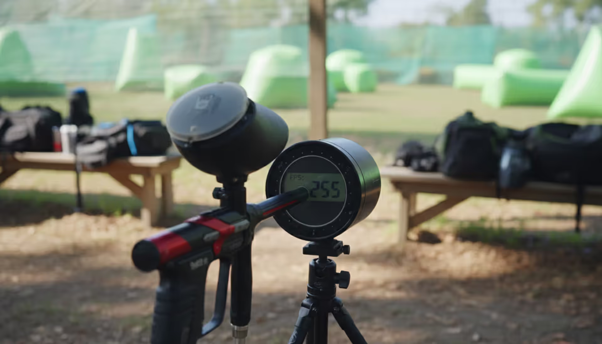 Paintball chronograph mounted on tripod at outdoor staging area with marker aimed at sensor and inflatable bunkers in background
